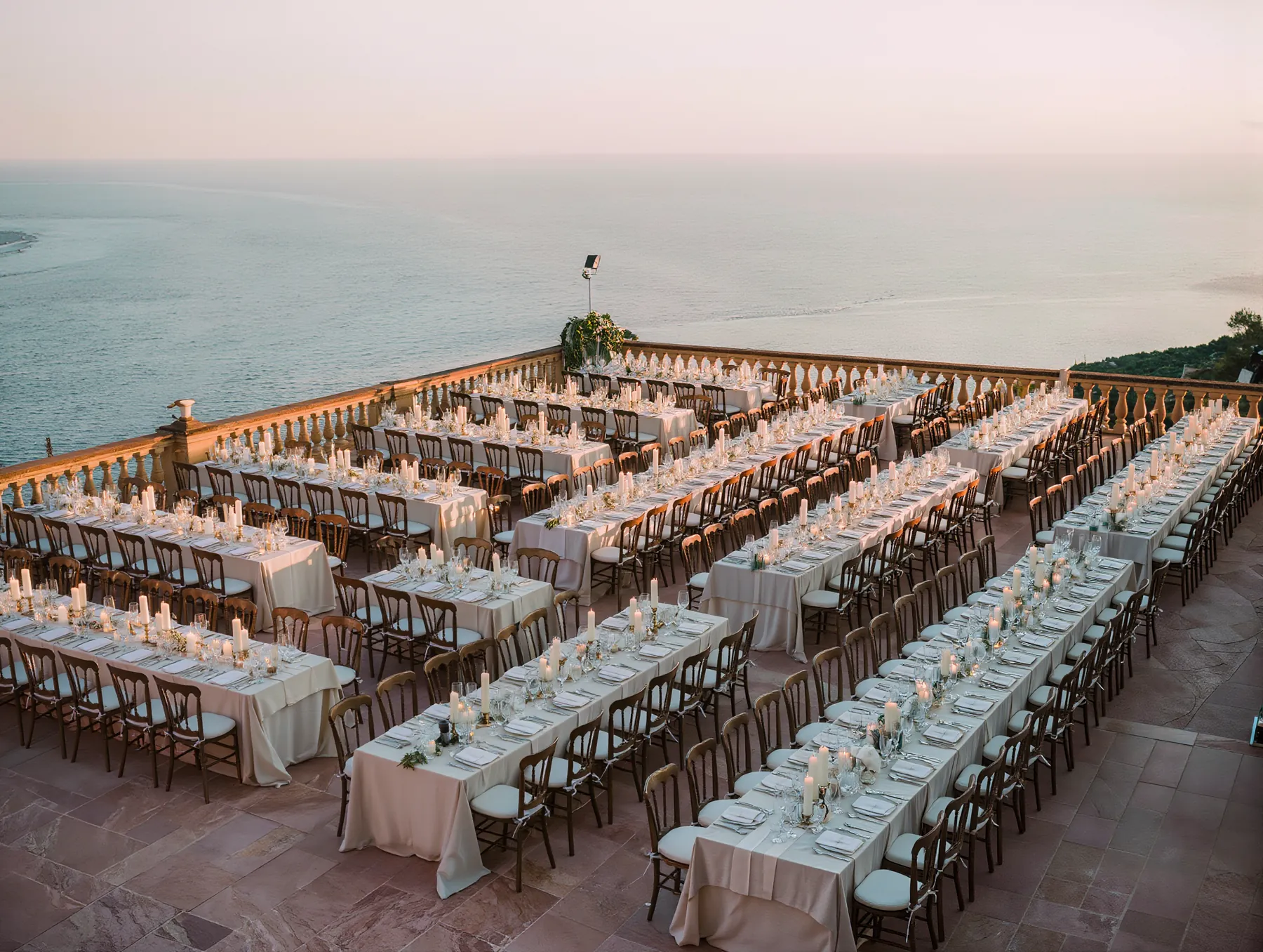 Long banquet tables laid with candles and glassware across a stone terrace above the sea, set for a large evening gathering.