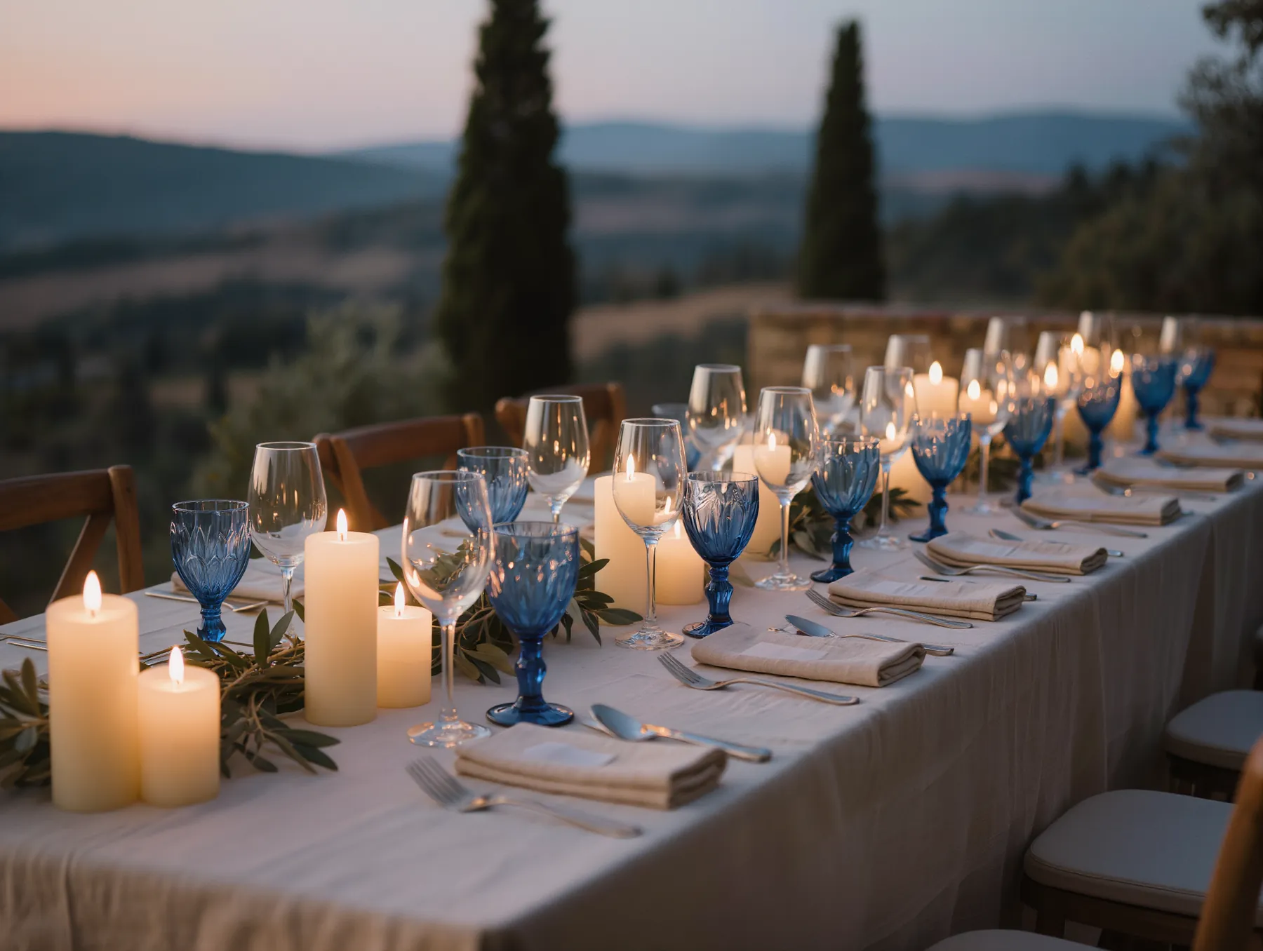 A candlelit dinner table at dusk with deep blue glassware, olive sprigs and linen napkins, overlooking hills and cypress trees.
