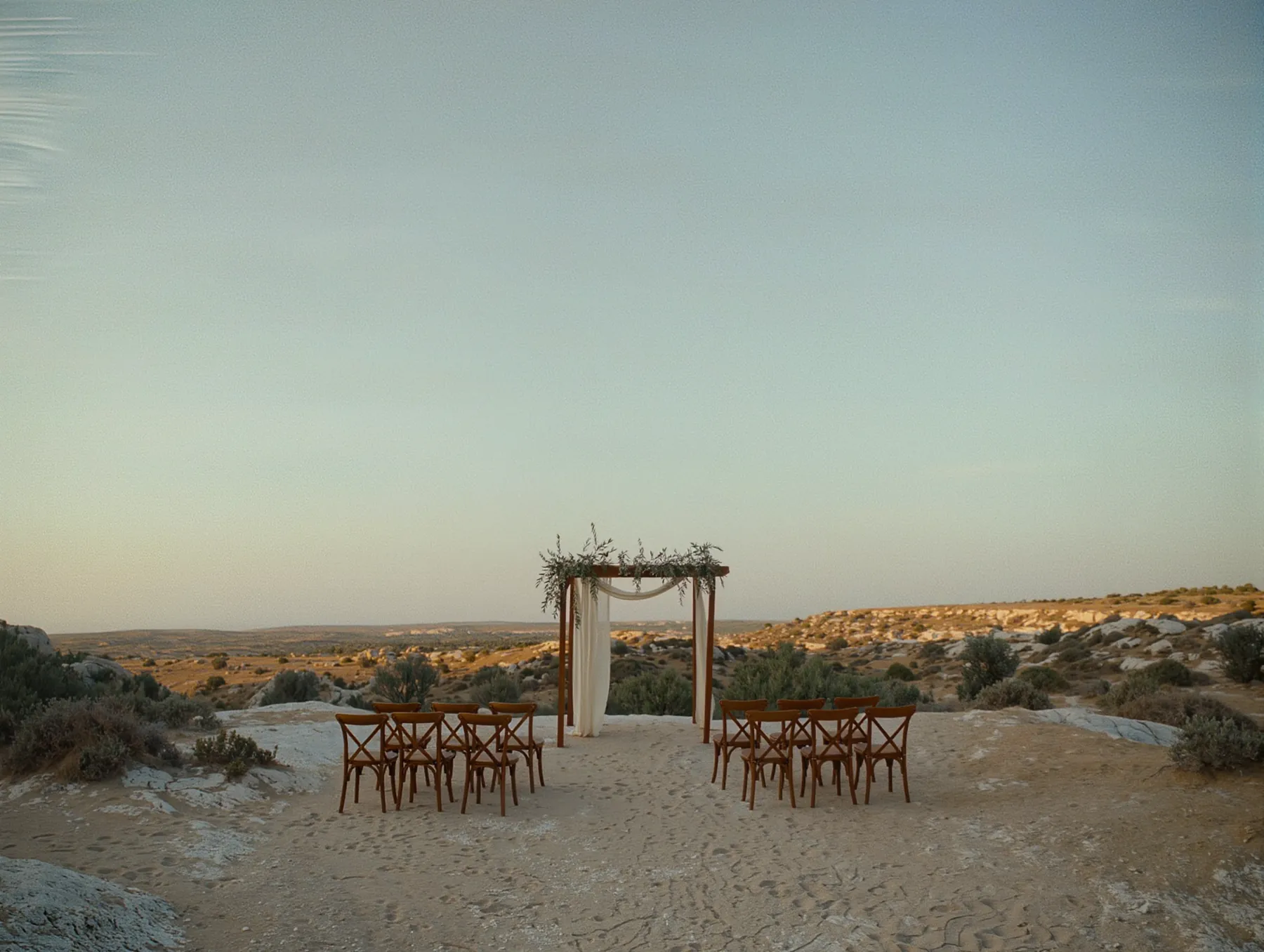 A ceremony set on a clifftop at dusk: a timber arch dressed with olive branches and linen drapes, two short rows of crossback chairs on pale rock.