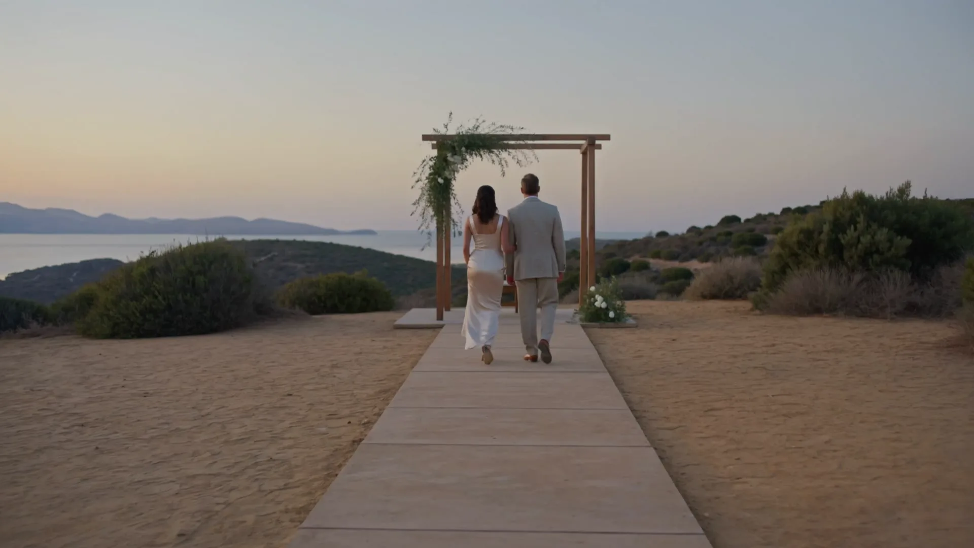 A couple walking hand in hand toward an olive-draped altar at dusk, seen from behind.